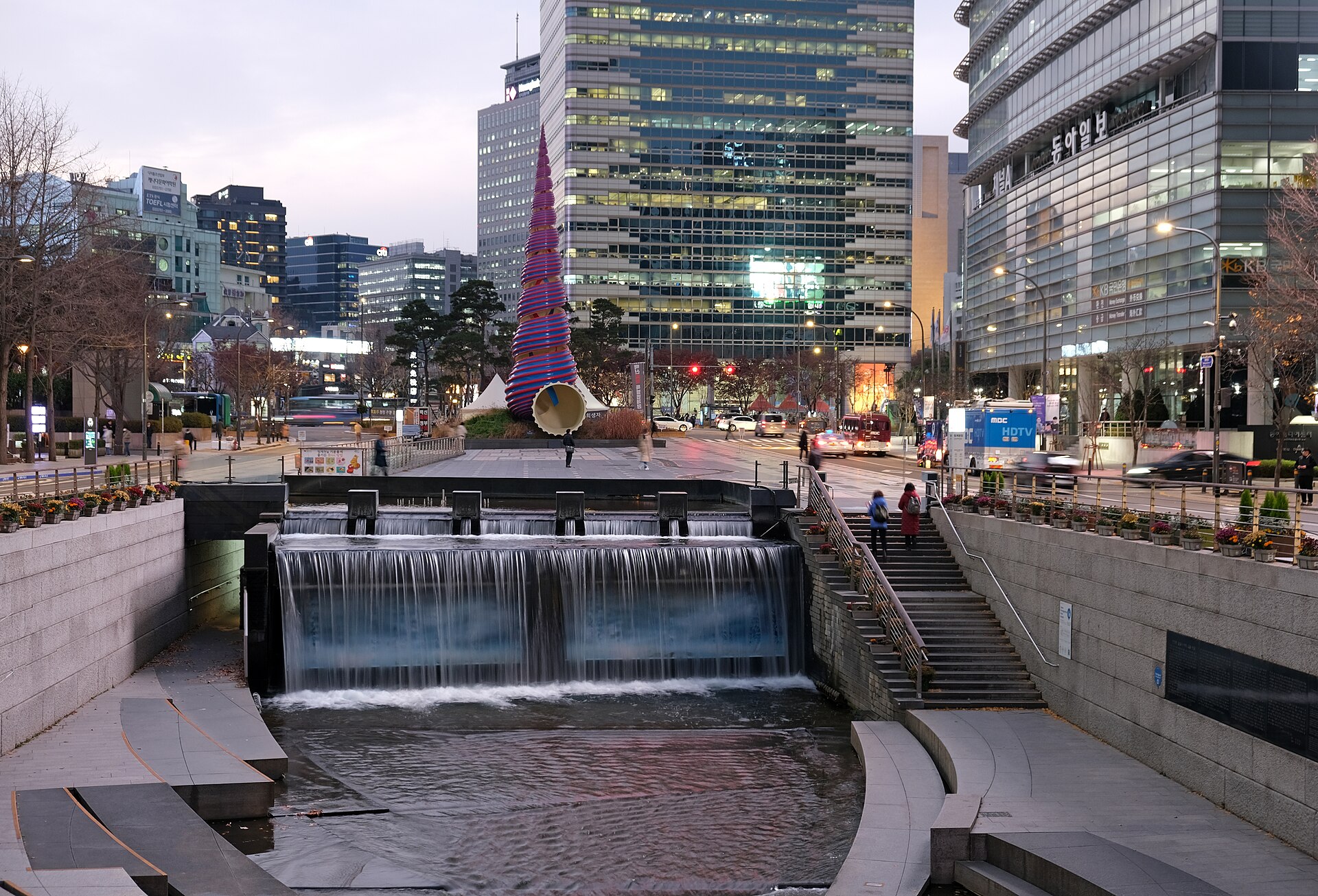 Thác nước đầu nguồn Cheonggyecheon buổi tối với bậc ngồi ven suối và tháp điêu khắc xoắn màu hồng xanh