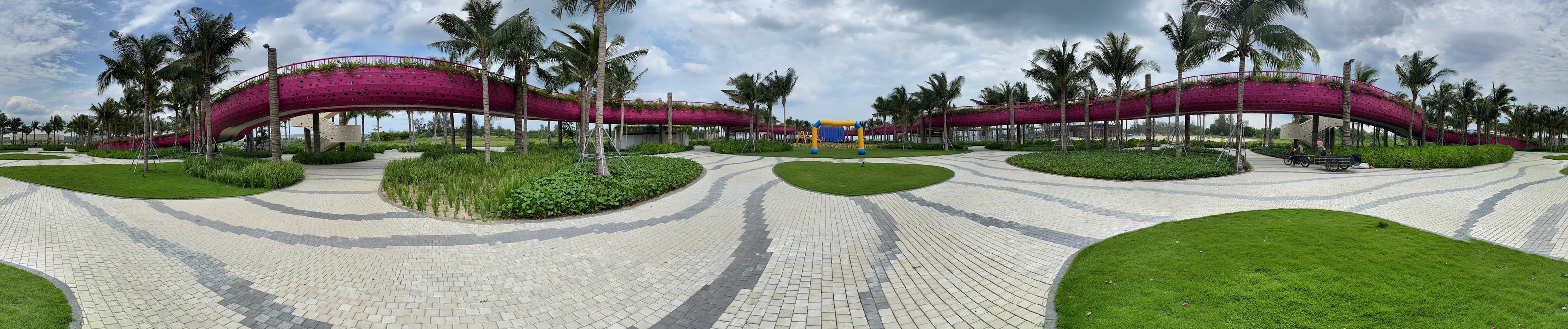 Ảnh toàn cảnh 360 độ trải phẳng của quảng trường Thanh Long Baywalk: tuyến cầu đi bộ màu hồng sen uốn quanh, hàng dừa, nền lát đá sáng màu vẽ những dải vân sóng, thảm cỏ bo tròn