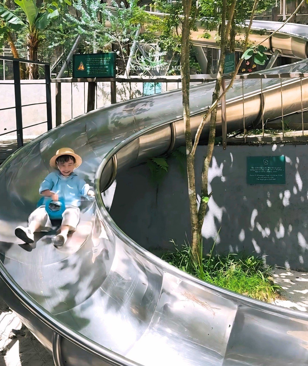 Một bé đội mũ cói ngồi trong lòng máng trượt ống inox uốn cong, nắng lọt qua tán thông in đốm trên mặt thép