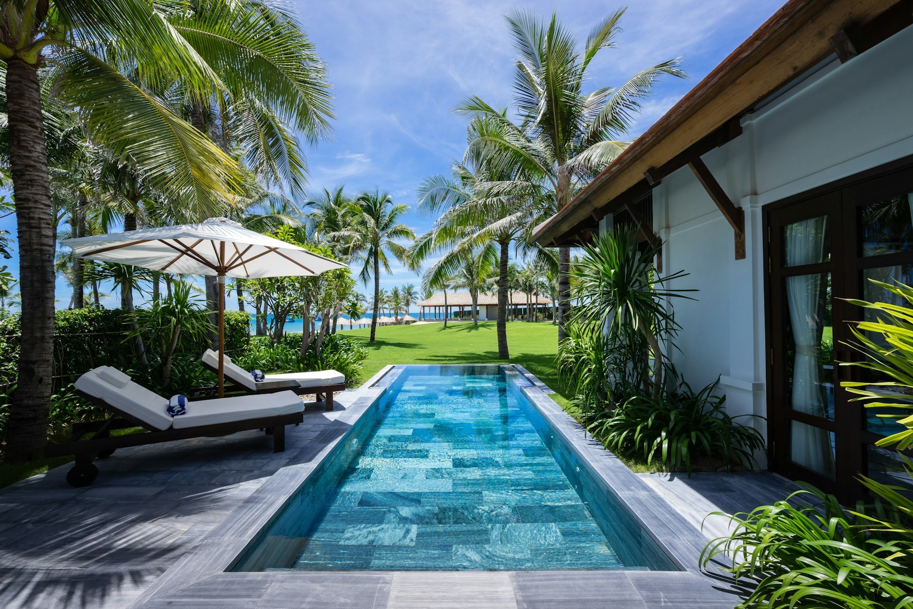 Sân sau một căn villa: hồ bơi dài hẹp, ghế nằm, dù trắng, phía xa là mái tranh và biển
