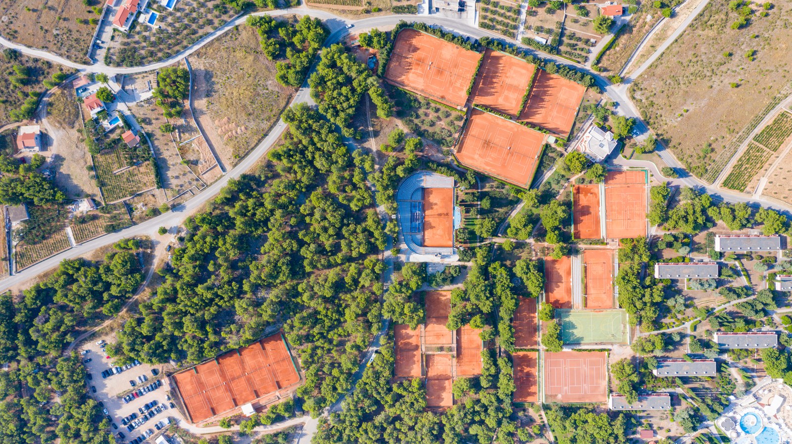Ảnh chụp thẳng đứng từ trên xuống một khu nghỉ dưỡng thể thao: hơn mười sân tennis đất nện màu cam nằm rải giữa rừng thông, một sân trung tâm có khán đài, và các khối lưu trú thấp tầng ở mép phải khung hình