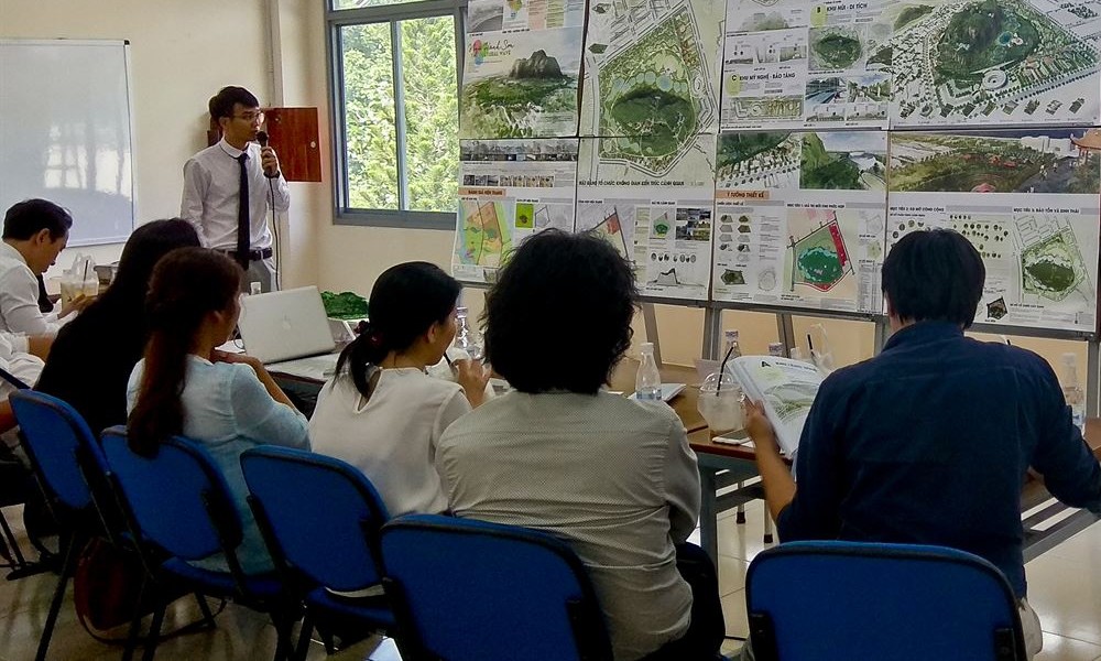 Người trình bày cầm micro, thành viên hội đồng đọc thuyết minh in sẵn trước giá bản vẽ