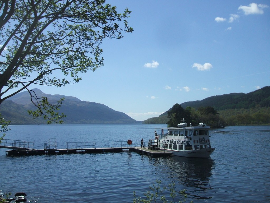 Tàu du lịch trắng mang dòng chữ Cruise Loch Lomond cập cầu tàu nổi, núi và rừng cây phía sau