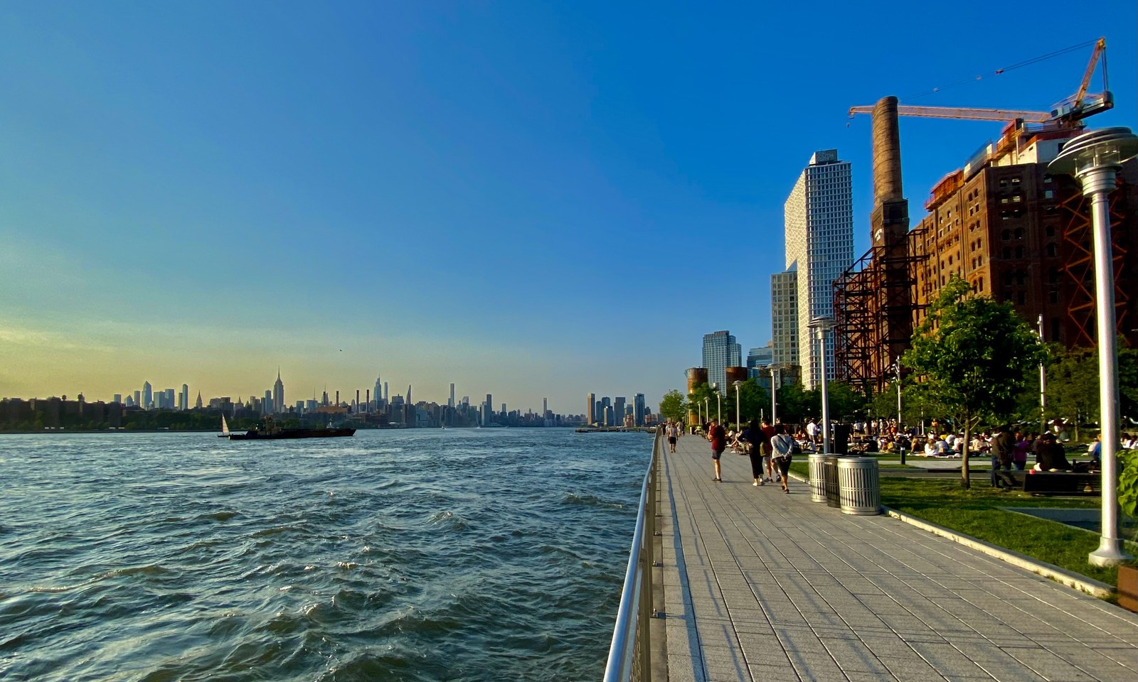 Lối đi ven sông với hàng ghế, bãi cỏ và đường ray cũ, phía xa là đường chân trời Manhattan