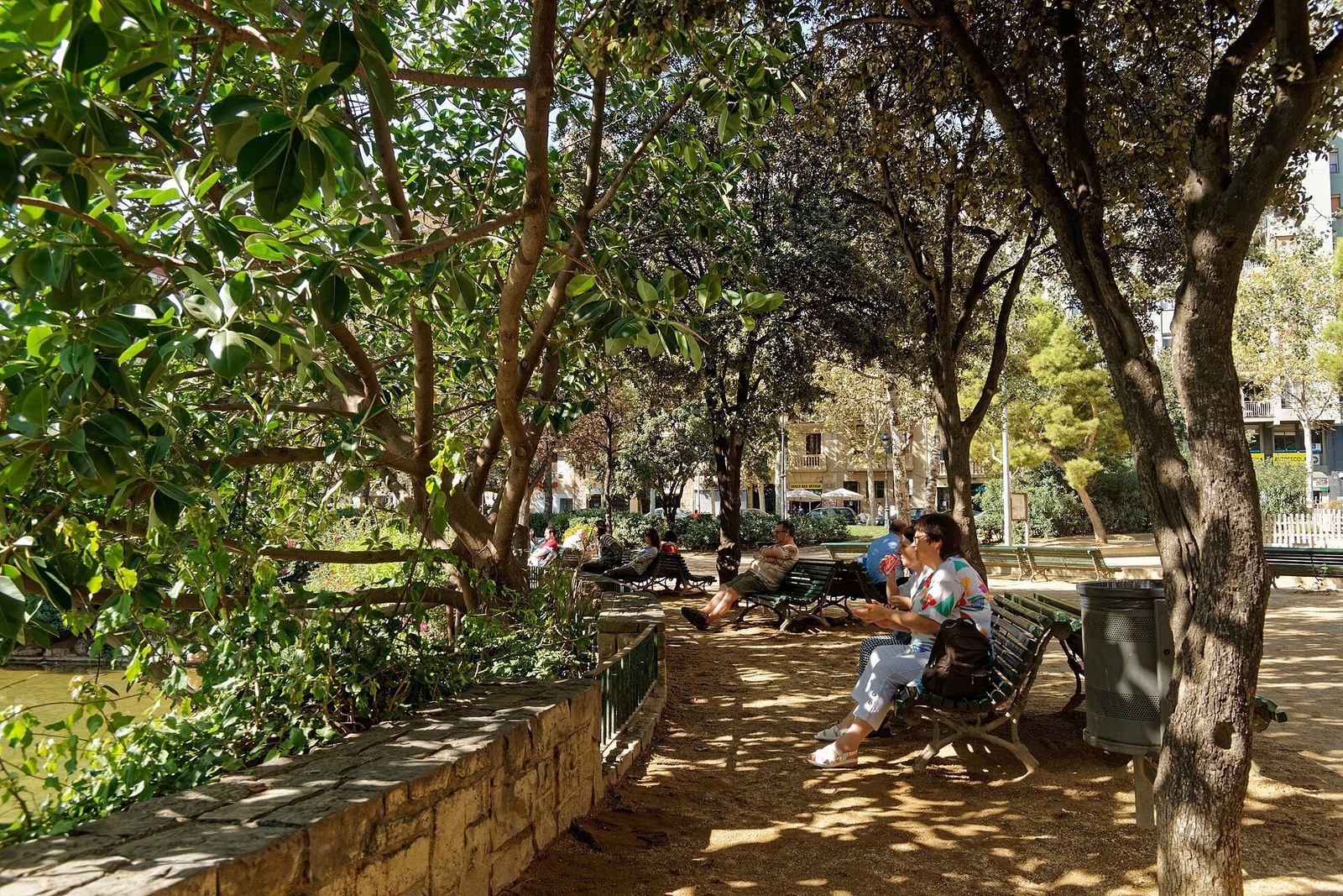 Sagrada Família — lối dạo nền đất dưới tán cây ở Plaça de Gaudí, người ngồi ghế cạnh bờ tường hồ