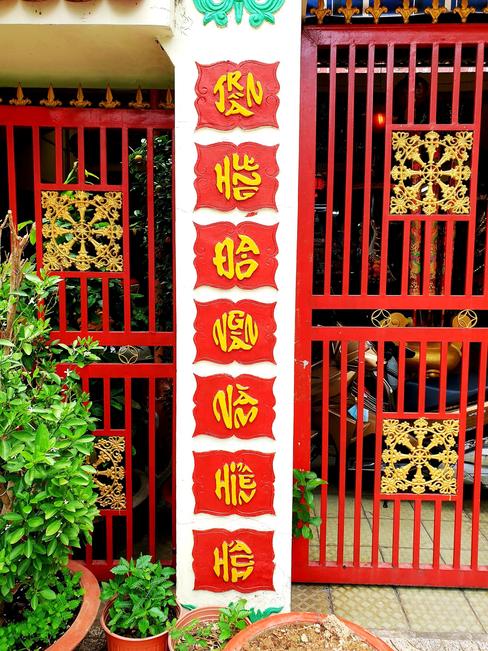 Trụ cổng trắng gắn dải câu đối đỏ chữ vàng cạnh cửa sắt đỏ