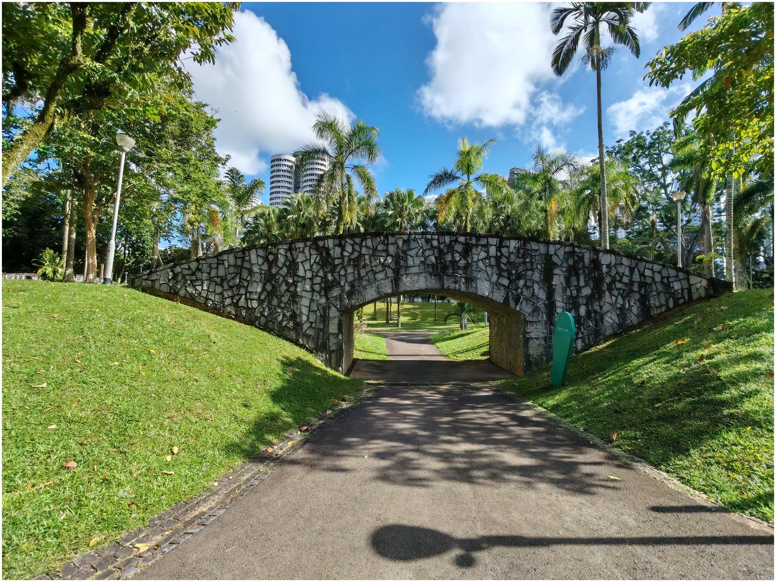 Bishan–Ang Mo Kio Park, Singapore — cây cầu đá kiểu vòm bắc qua lối đi bộ, hai bên trồng cọ, nhà cao tầng phía xa