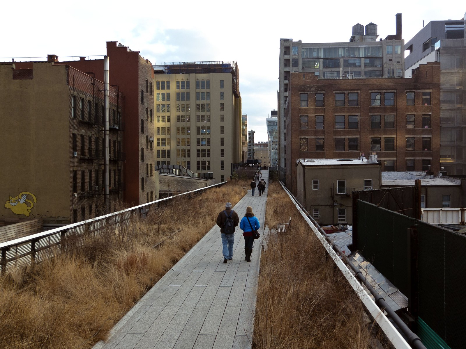 High Line — Lối đi lát tấm bê tông giữa dải cỏ khô cuối đông, hai bên là nhà gạch cũ