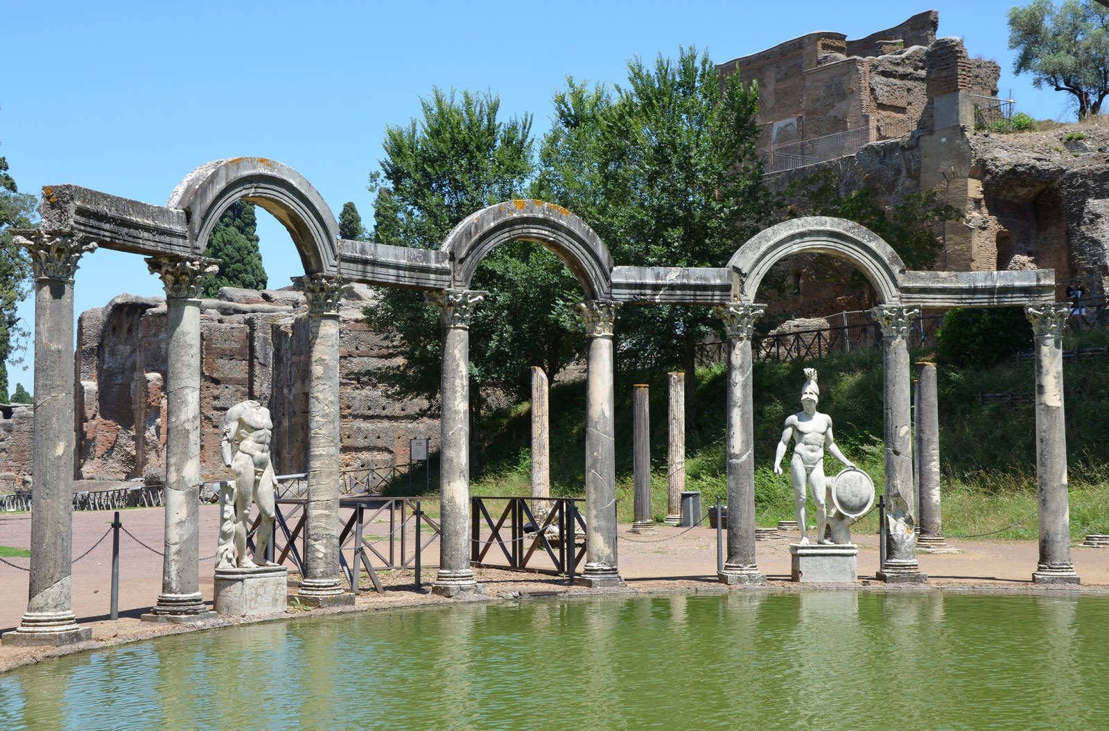 Dãy cột đá cong hình vòm cuốn ven một hồ nước dài tại Canopus, Hadrian's Villa, hai tượng đá trắng đứng cạnh hồ và tàn tích gạch phía sau