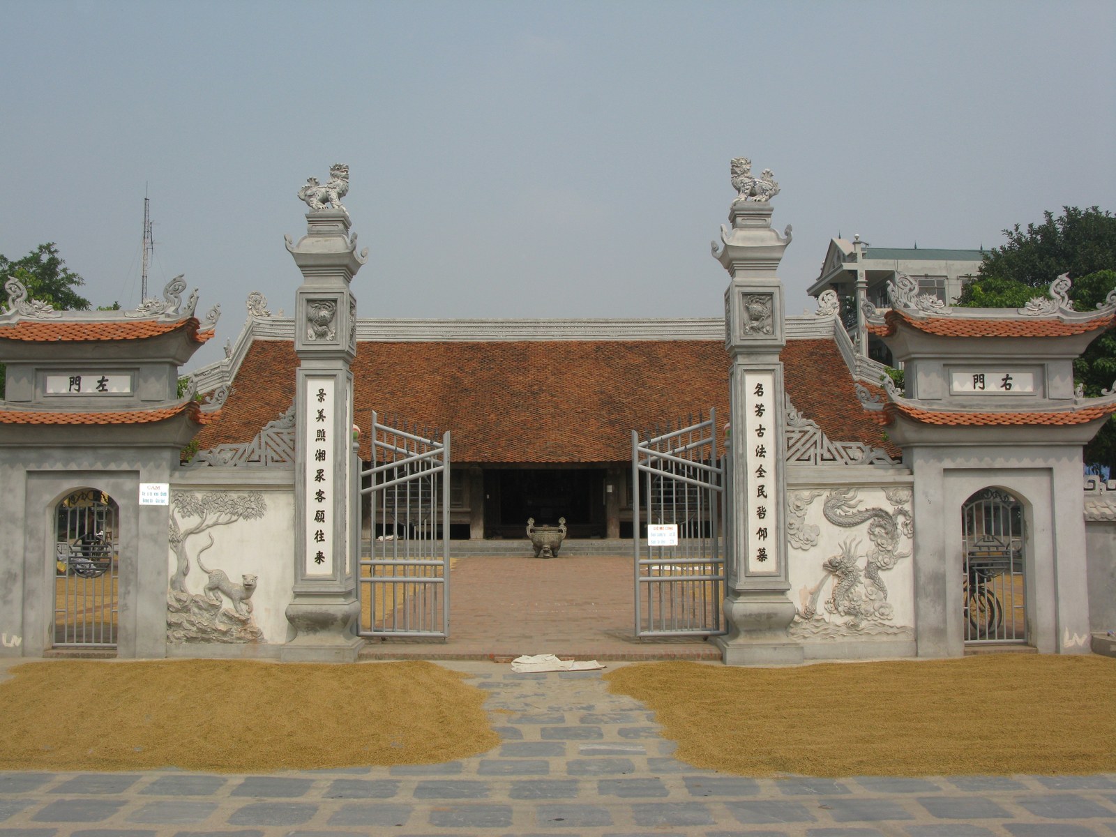 Cổng đình Đình Bảng nhìn thẳng: hai trụ cổng lớn có tượng lân, cổng sắt mở, phía sau là mái đình cong đỏ, sân phơi thóc