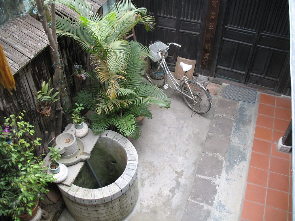 Sân trong nhỏ của một nhà cổ, nhìn từ trên xuống: bể nước xây gạch tròn, cây cau cảnh, một xe đạp dựng cạnh cửa gỗ đen