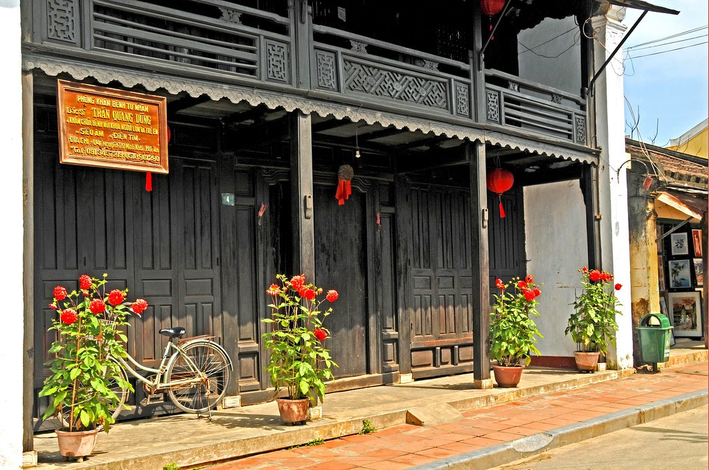 Nhà cổ Phùng Hưng, Hội An: mặt tiền gỗ sơn đen hai tầng, ban công chạm khắc hoa văn hình quả trám, chậu hoa đỏ đặt trước cửa