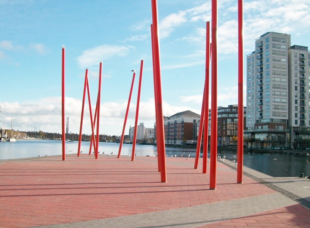 Hàng cột thép sơn đỏ dựng nghiêng trên quảng trường lát đá ven bến cảng Dublin
