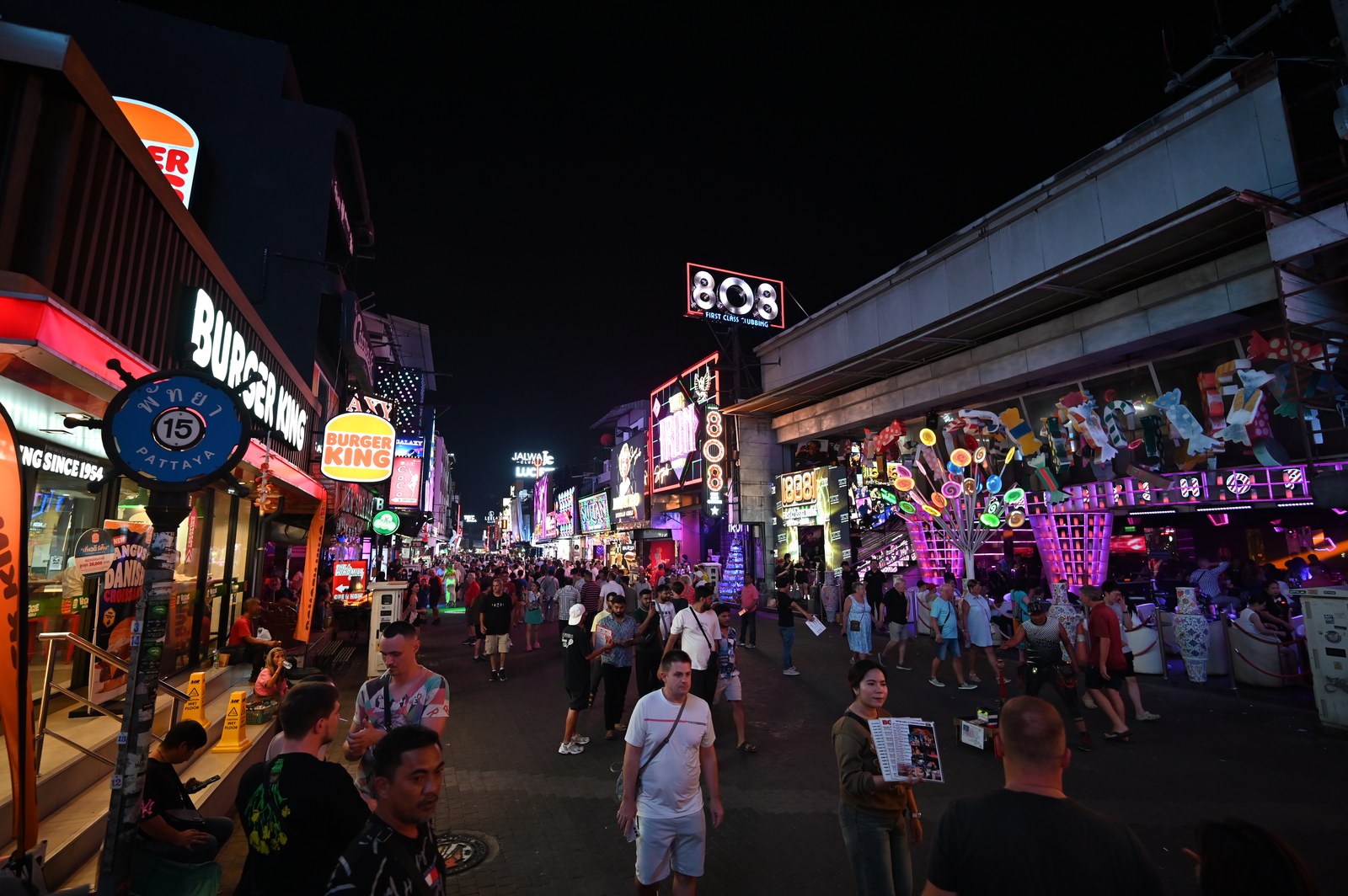 Một phố đi bộ về đêm rực rỡ biển hiệu neon của các quán bar, cửa hàng đồ ăn nhanh và câu lạc bộ, đông người đi lại giữa lòng phố.