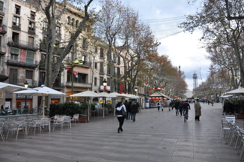 Đại lộ La Rambla có hàng cây hai bên, bàn ghế cà phê ngoài trời xếp sát các ki-ốt ven đường, người đi bộ và một người đi xe đạp ở giữa lối đi lát đá.