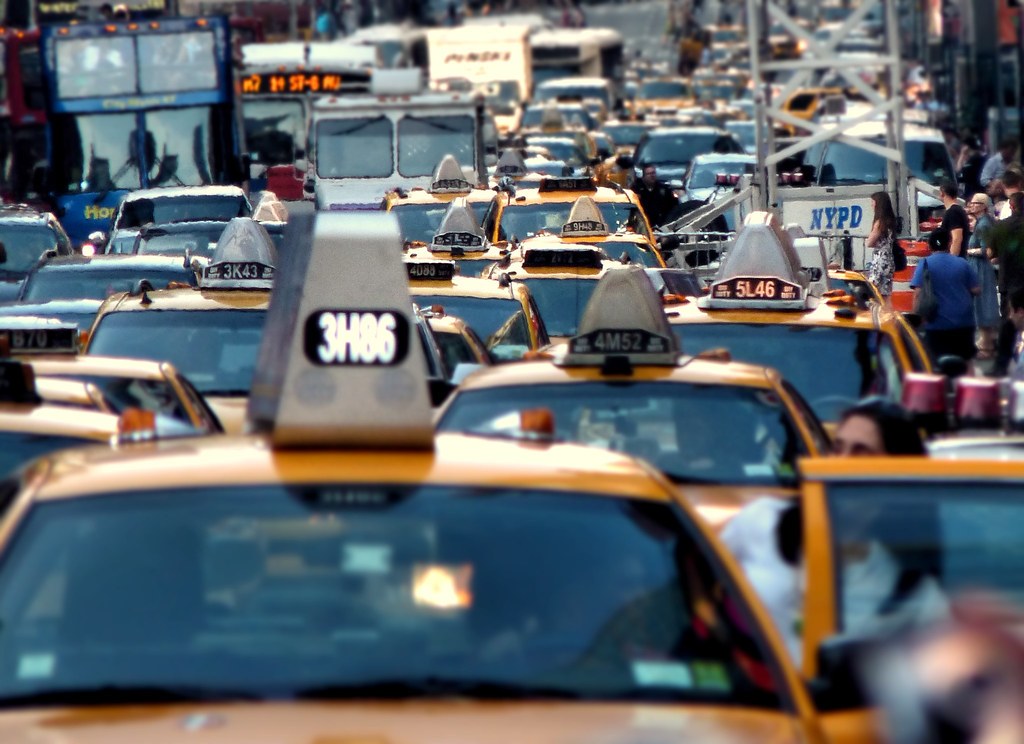 Rất nhiều taxi màu vàng xếp sát nhau kín cả làn đường, một xe cảnh sát NYPD và vài người đi bộ len giữa dòng xe.