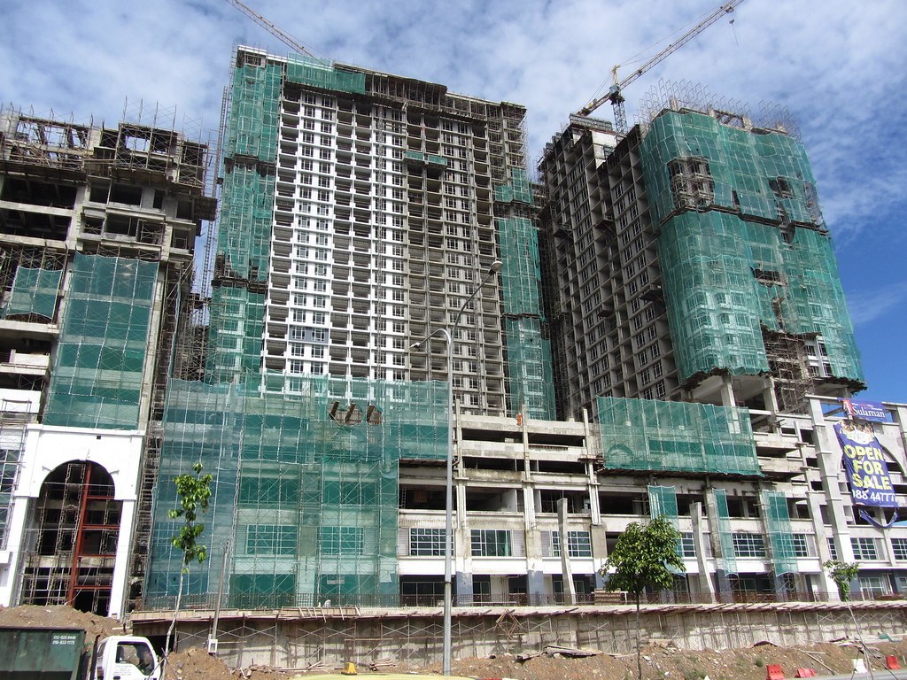 Toà tháp cao tầng đang xây, bọc lưới an toàn xanh, hai cần trục phía trên, biển quảng cáo bán căn hộ ở góc phải